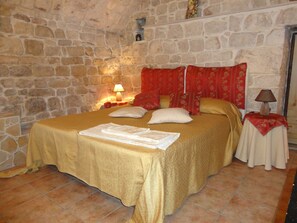 Ein Zimmer mit einer Steinwand, einem Bett mit goldener Bettdecke, zwei Kissen, einem roten Kopfteil und zwei Nachttischen mit Lampen.