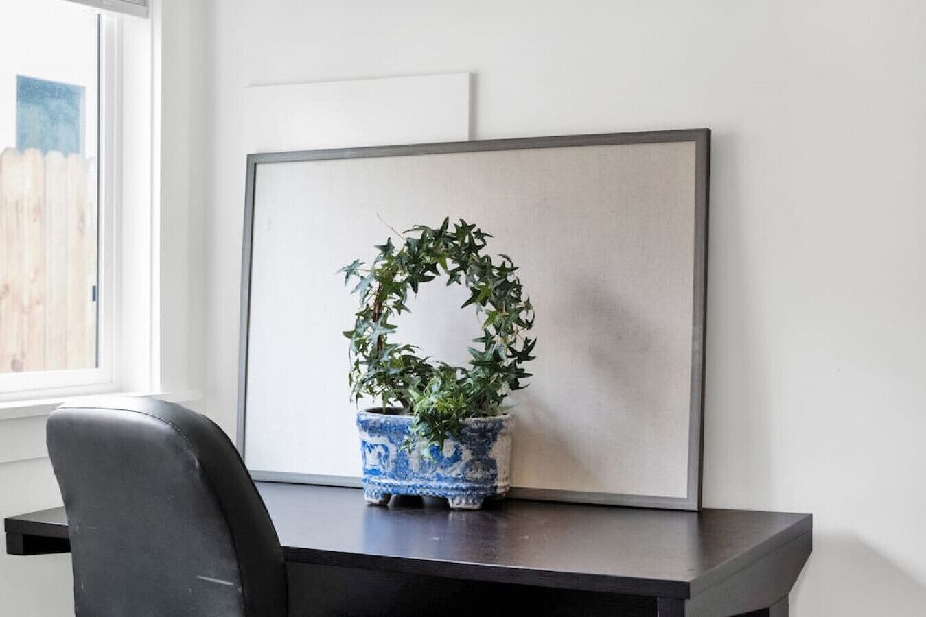 Un espace de travail avec une plante en pot sur un bureau, un tableau encadré au mur et une fenêtre avec des stores.