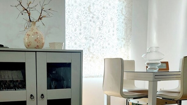 A dining area with a white table, two chairs, a vase with branches, and a patterned window blind.