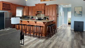 A kitchen with wooden cabinets, a central island with stools, a refrigerator, and hanging pendant lights. - Stay in Comfort at Corky's Cove Unit #1. (Port Mansfield)