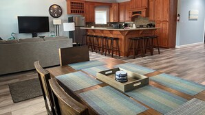 A modern kitchen with wooden cabinets, a central island with bar stools, a flat-screen TV, and a dining area with a wooden table and chairs. - Stay in Comfort at Corky's Cove Unit #1. (Port Mansfield)