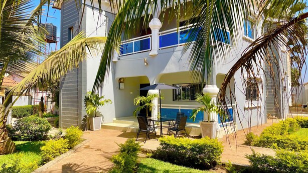 A two-story building with a balcony, a pool area, and a garden with palm trees. - The Unique and Tranquil Retreat Apartment (Brufut)