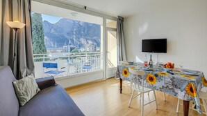 Une pièce comprenant un canapé, une table à manger recouverte d’une nappe à motifs de tournesols, un téléviseur et un balcon offrant une vue sur la montagne.