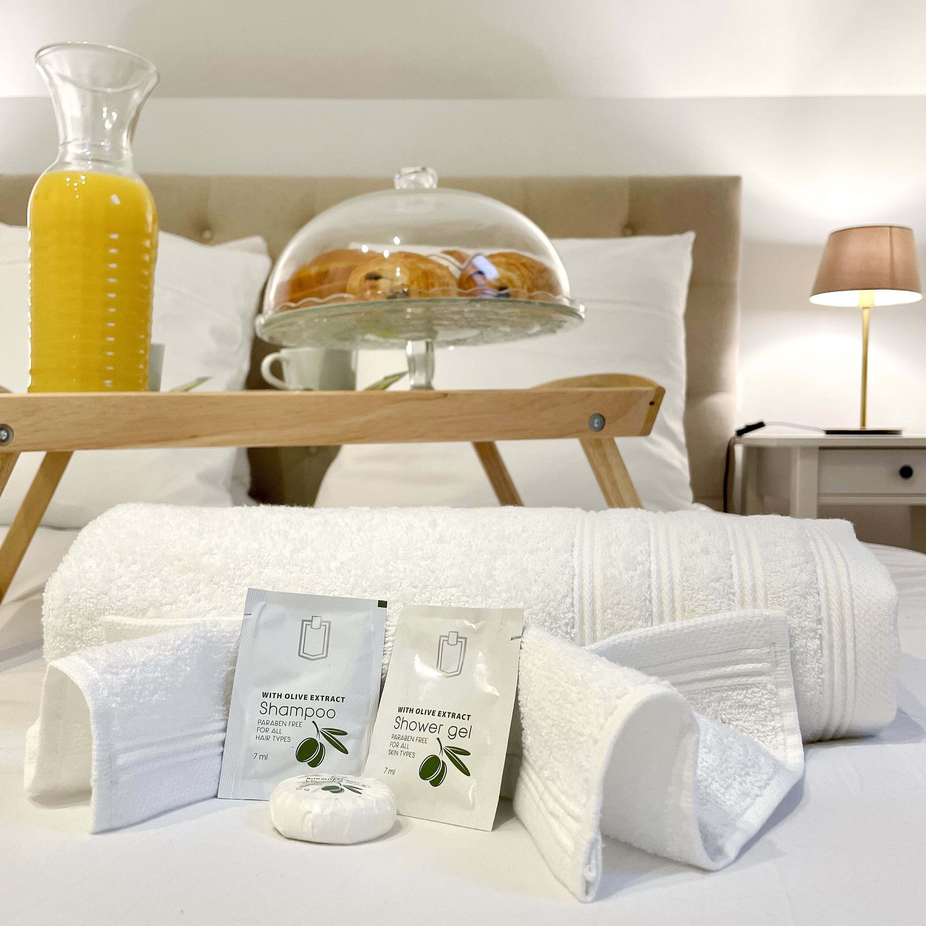 A neatly made bed with white towels, a glass bottle of orange juice, a glass of water, a shower gel package, and a shampoo package.