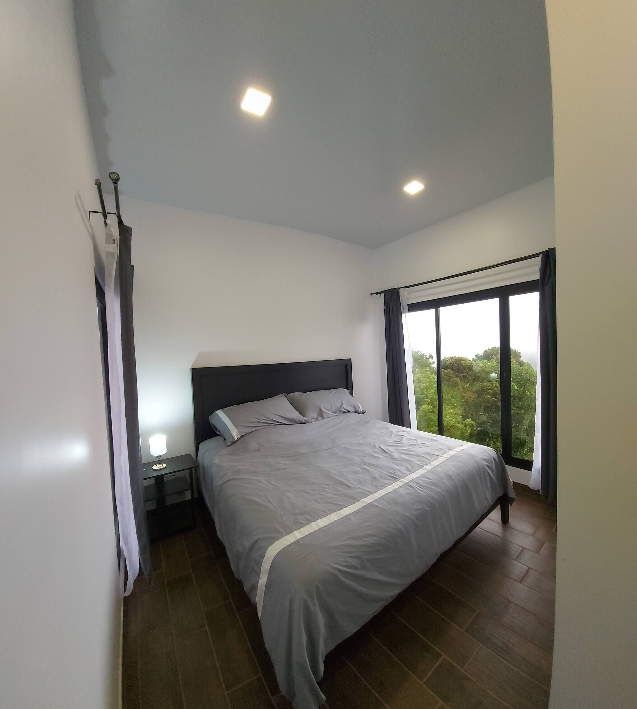 A bedroom with a bed, a nightstand, a window with curtains, and a view of greenery outside.