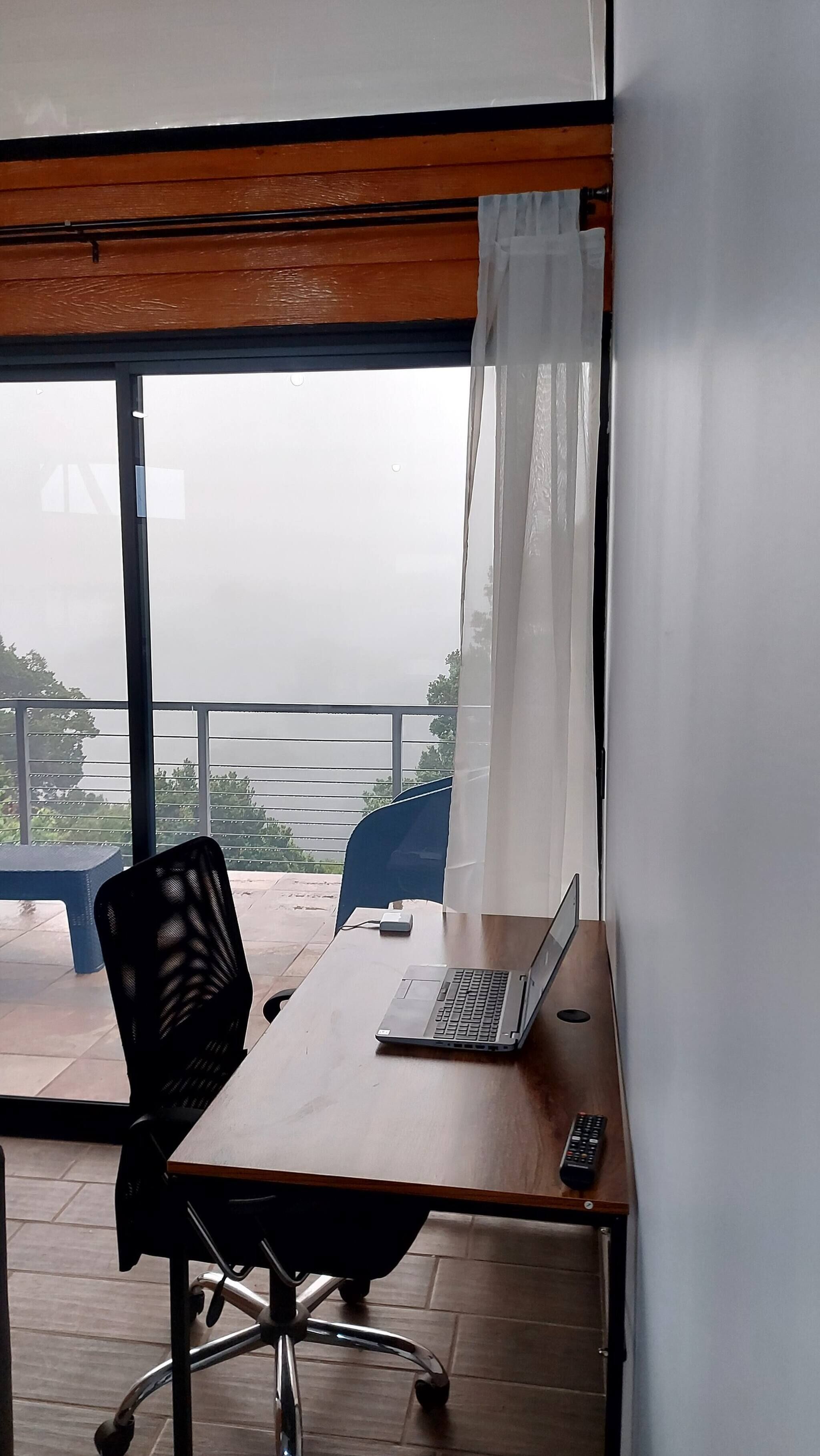 Quarto com escrivaninha de madeira, laptop, cadeira de escritório preta, janela com vista para uma varanda e cortina.