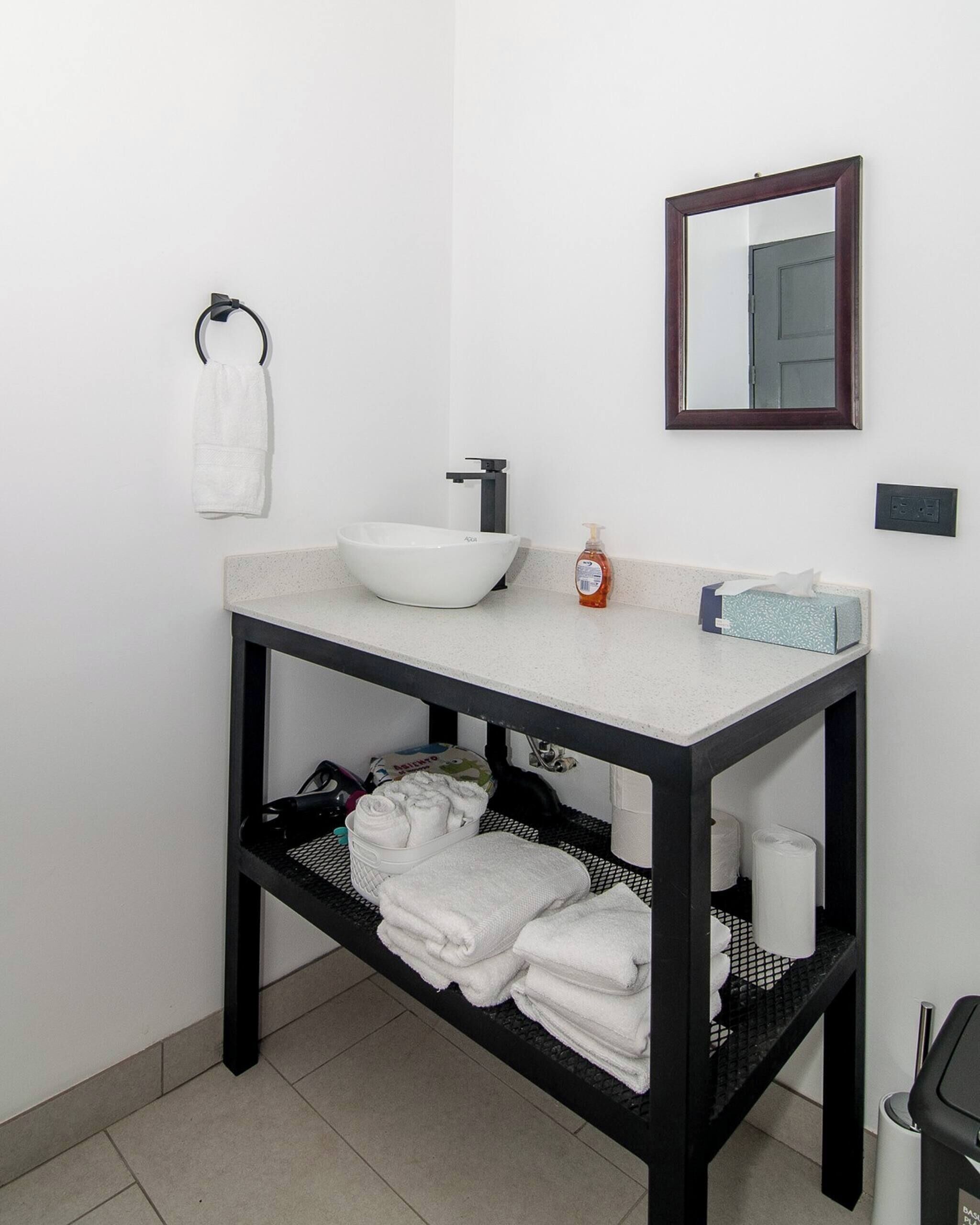 Een badkamer met een witte wastafel, een spiegel, een handdoekhouder, een zeepdispenser en een plank met handdoeken en toiletartikelen.