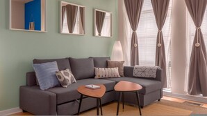 Ein Wohnzimmer mit grauem Sofa, zwei Beistelltischen und einer Lampe. An der Wand befinden sich drei Spiegel und ein Fenster mit Vorhängen.