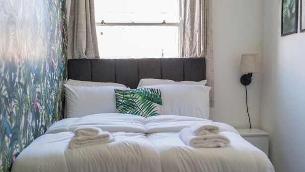 A neatly made bed with white linens and a green leaf pillow, a bedside lamp, and a window with curtains. - Studio Unit in Chapel Market London (London)
