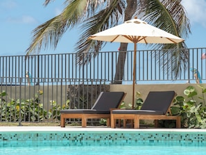 A poolside area with two lounge chairs and a beach umbrella, surrounded by a fence and palm trees. - Appartement Orient Bay-ti Paradise (Collectivité de Saint-Martin)