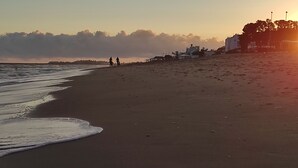 Ein Strand mit blauem Himmel, Wellen und einigen Spaziergängern. Im Hintergrund sind Gebäude zu sehen.