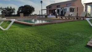 Ein Poolbereich mit Rasenfläche, Liegestühlen und einem Gebäude mit überdachter Terrasse.