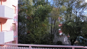 Un balcone con un tavolo apparecchiato per due persone, circondato da una ringhiera bianca, con vista su una zona residenziale con alberi e palazzi.