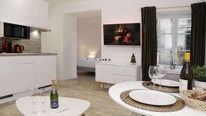 Eine moderne Küche mit weißen Schränken, einer Mikrowelle und einem roten Wasserkocher. Es gibt einen Essbereich mit einem weißen Tisch, zwei Weingläsern und einer Flasche Champagner. Ein Fernseher ist über der Küche an der Wand angebracht.