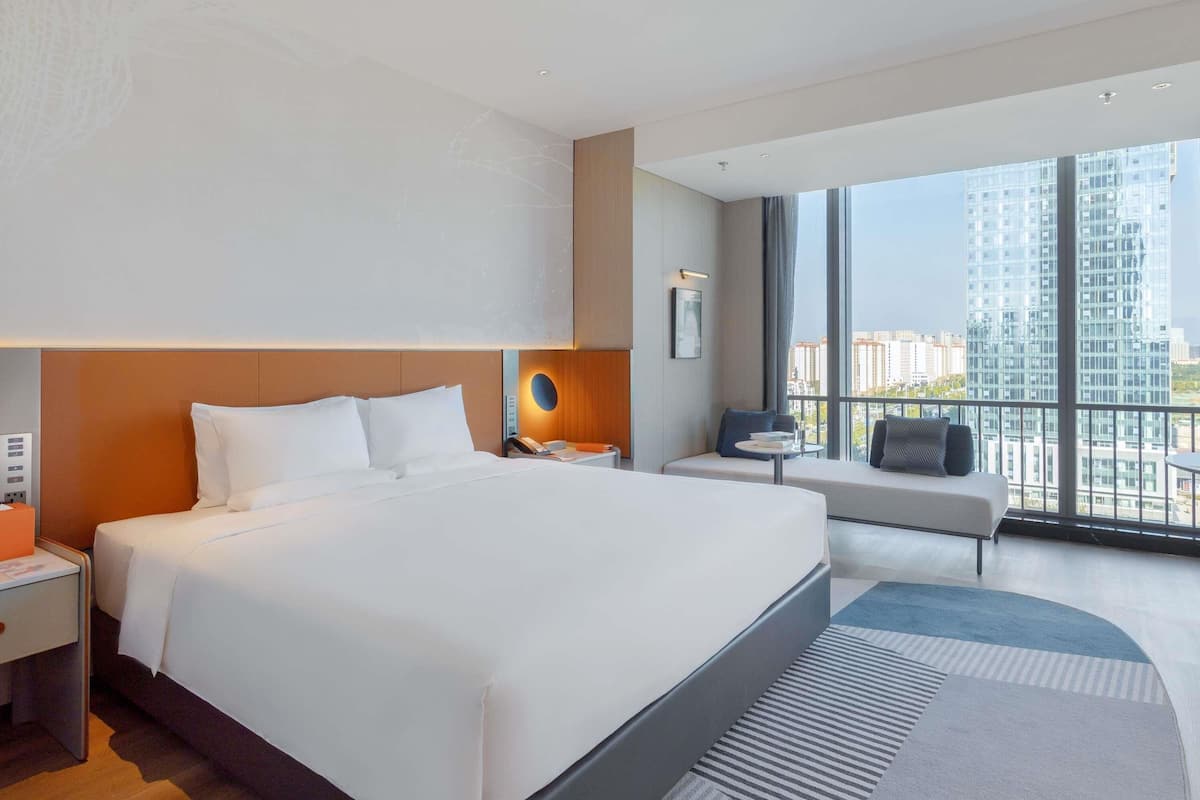Moderni hotellihuone, jossa on suuri sänky, yöpöytä lampun kanssa, pieni oleskelualue sohvalla sekä suuri ikkuna, josta avautuu näkymä kaupunkiin.