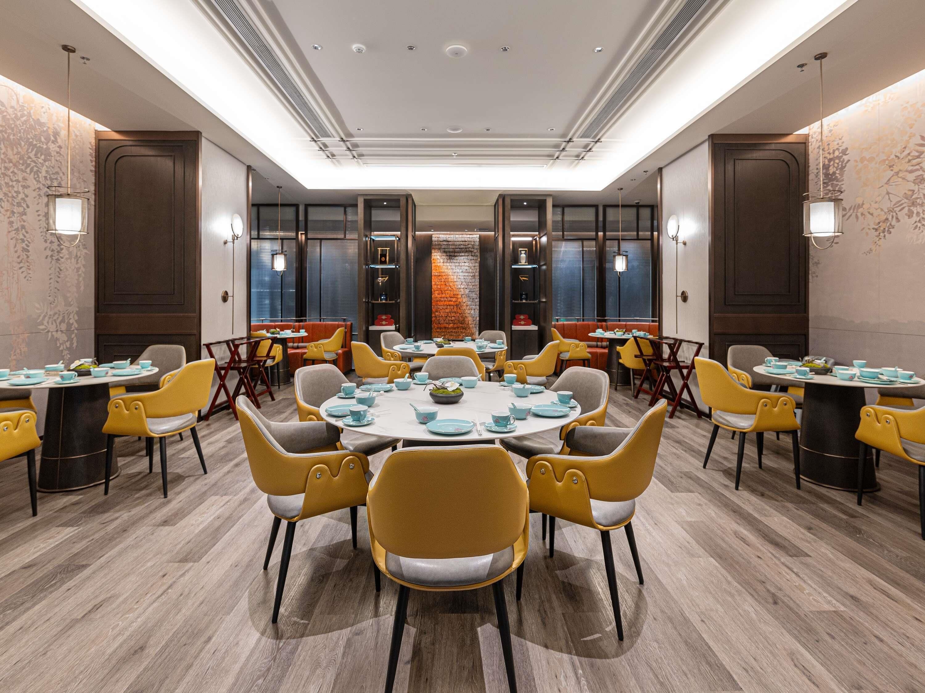 A spacious dining area with round tables and yellow chairs, featuring a modern design with wooden flooring and decorative wall panels.