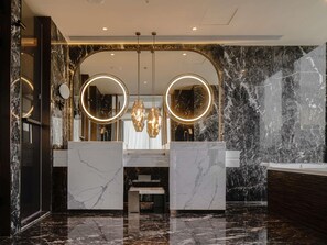 Un baño moderno con paredes de mármol, un lavamanos central y dos grandes espejos circulares con marcos iluminados.