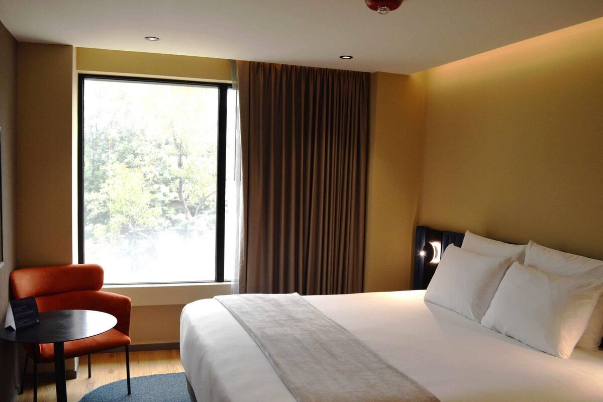 A hotel room with a bed, a chair, a table, and a window with curtains.