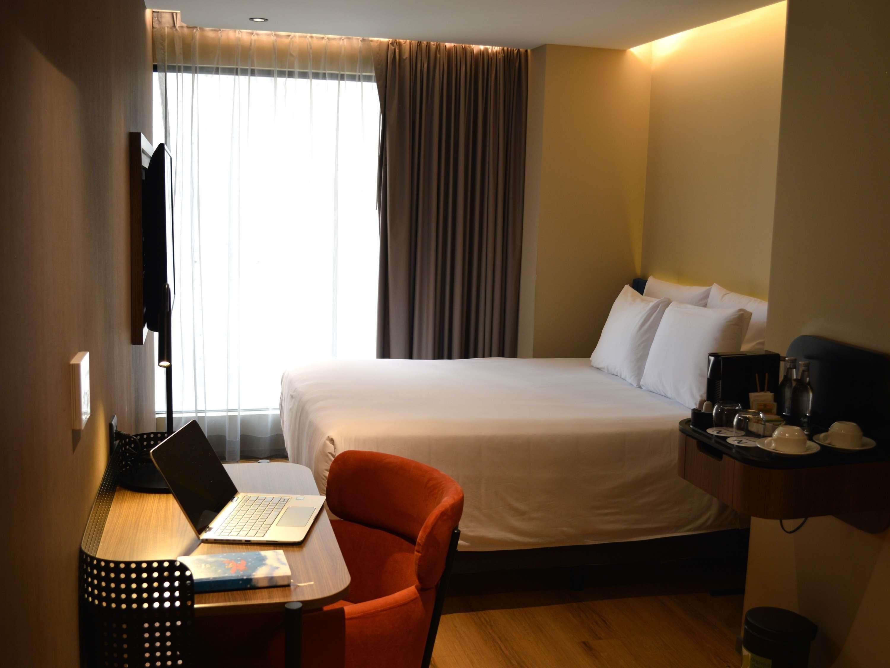 Habitació d'hotel amb un llit, una taula de treball amb un ordinador portàtil, una cadira, una tauleta de nit amb una cafetera i una finestra amb cortines.