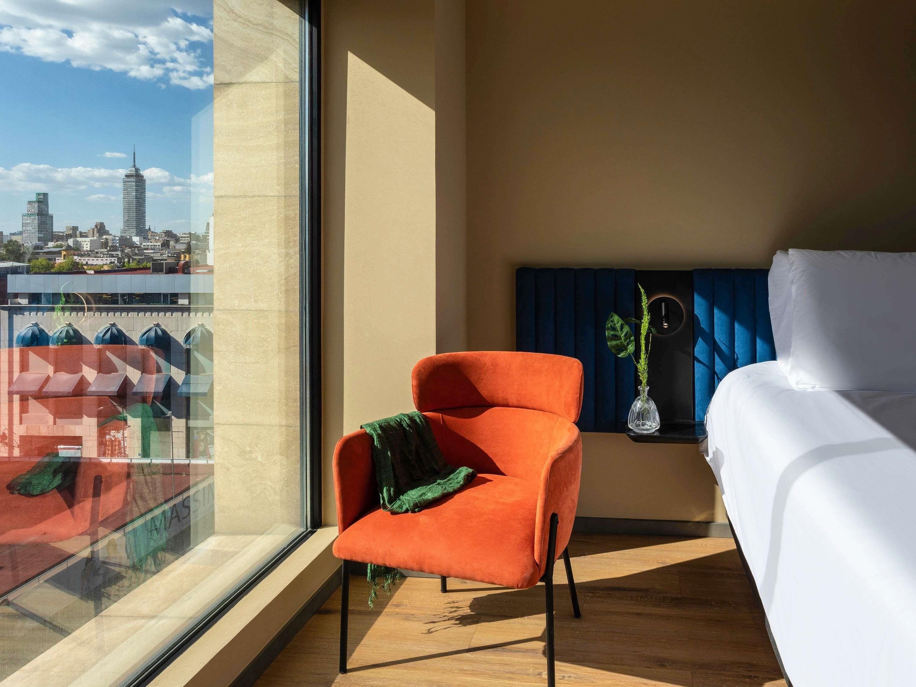 A hotel room with a large window, a bed, an orange chair, a nightstand with a lamp, and a view of a cityscape.