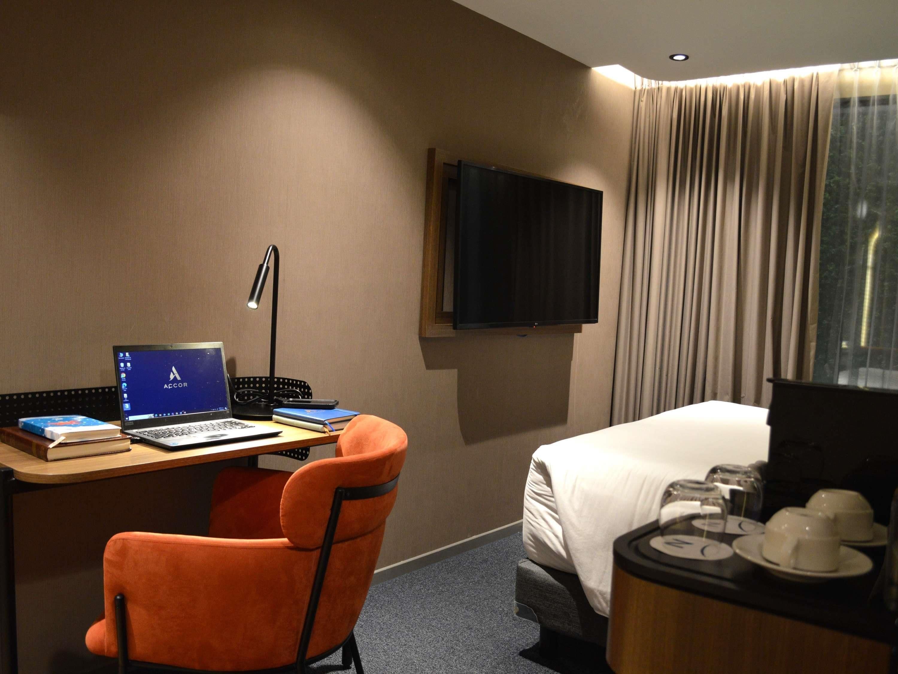 Une chambre d’hôtel équipée d’un bureau, d’un ordinateur portable, d’une chaise, d’un lit, d’une télévision et d’une fenêtre avec des rideaux.