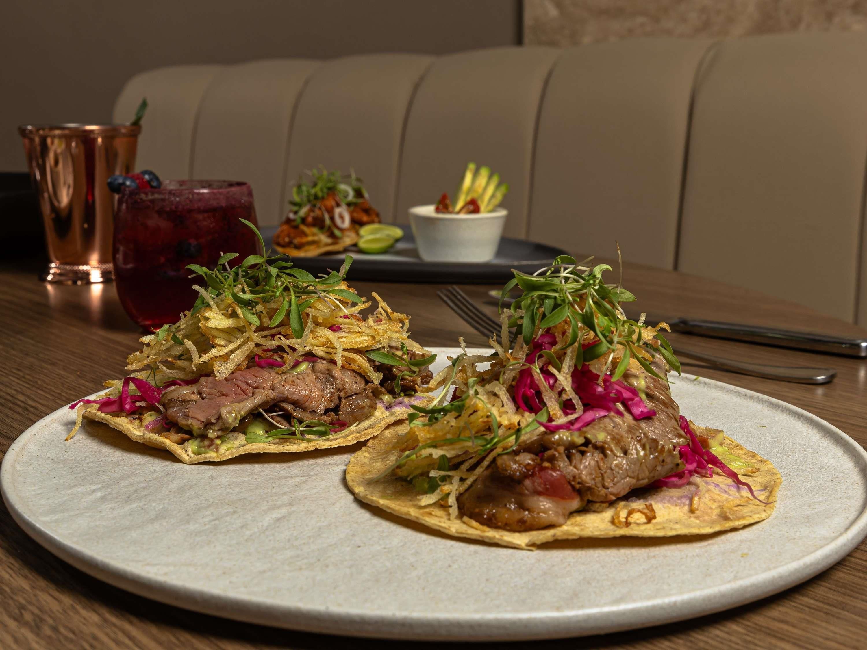 A plate of tacos with meat and vegetables on a ceramic plate, a glass of red drink, and a bowl of fries in the background.