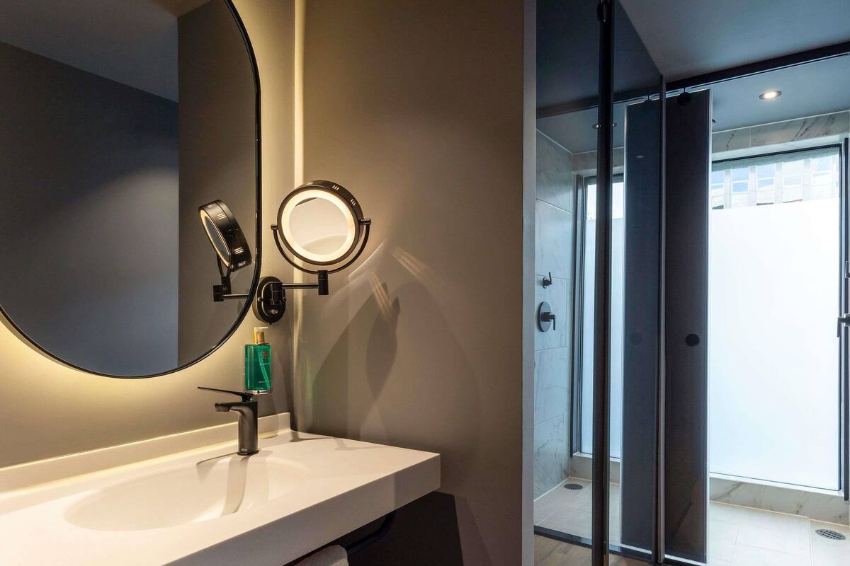 Un baño con un espejo grande, un espejo de tocador redondo, un lavabo y una puerta de ducha de cristal.