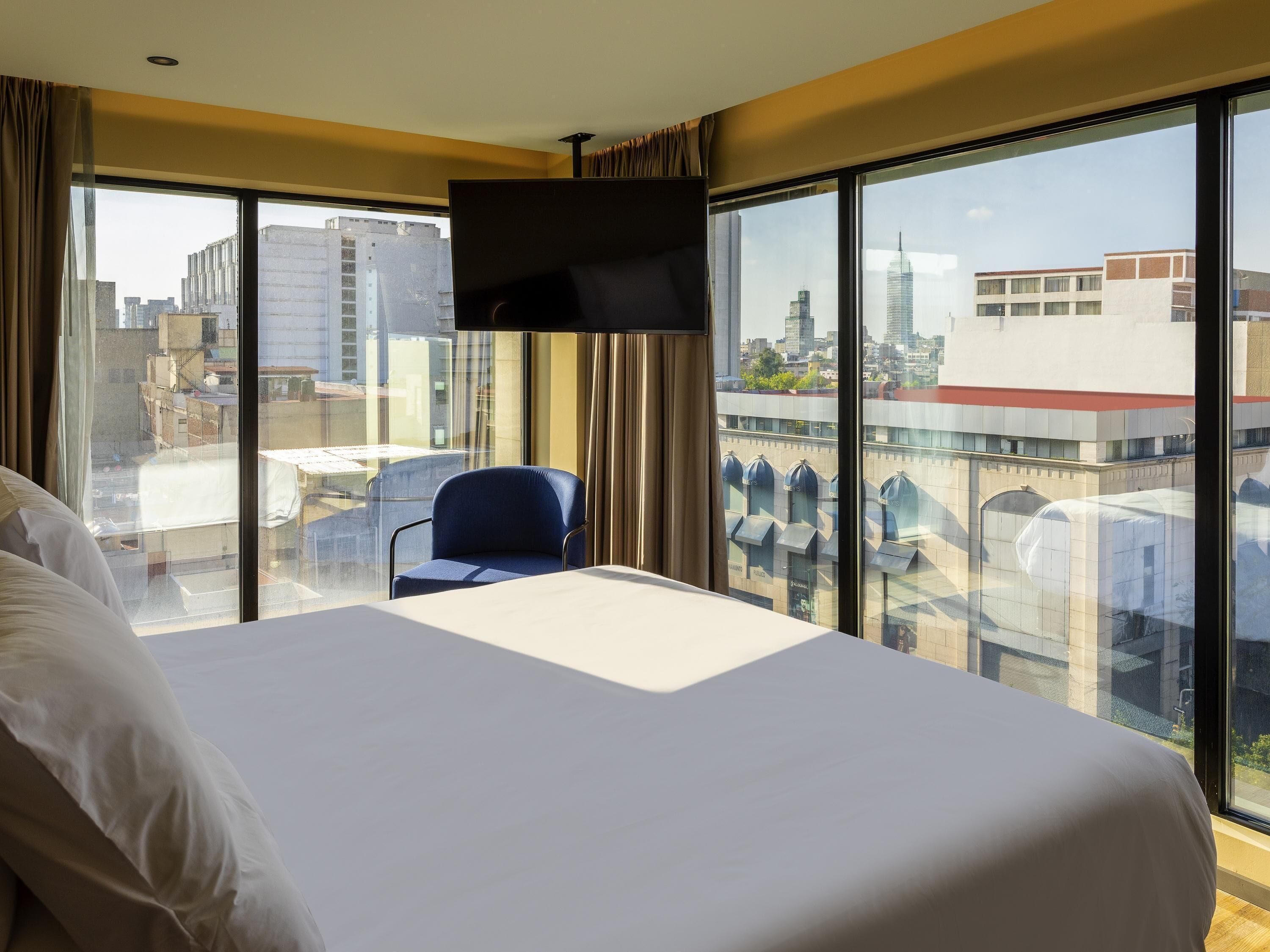 A hotel room with a large bed, a TV mounted on the wall, a chair, and a balcony with a city view.