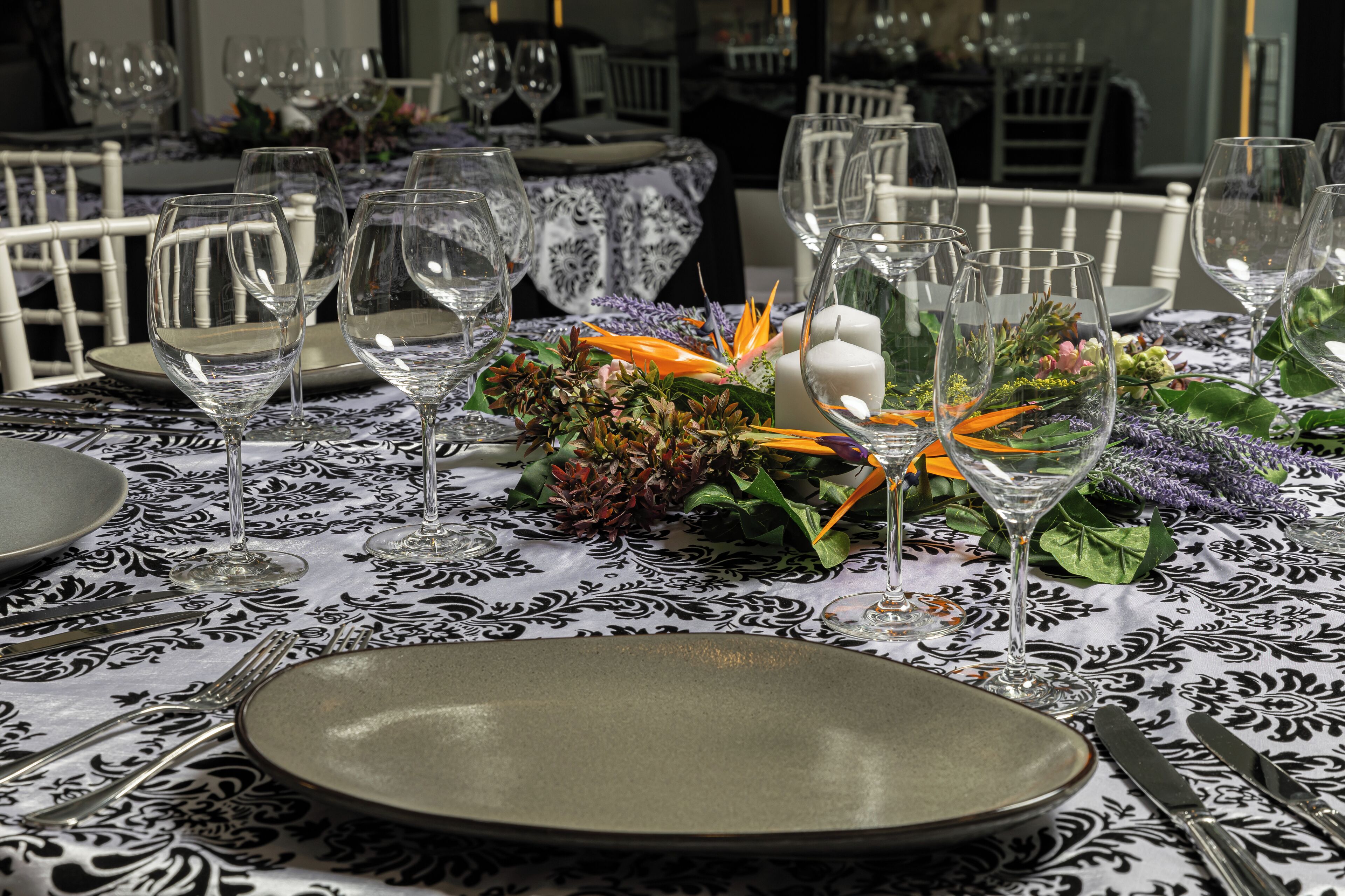 A dining table set with plates, cutlery, and wine glasses, surrounded by chairs and a patterned tablecloth.