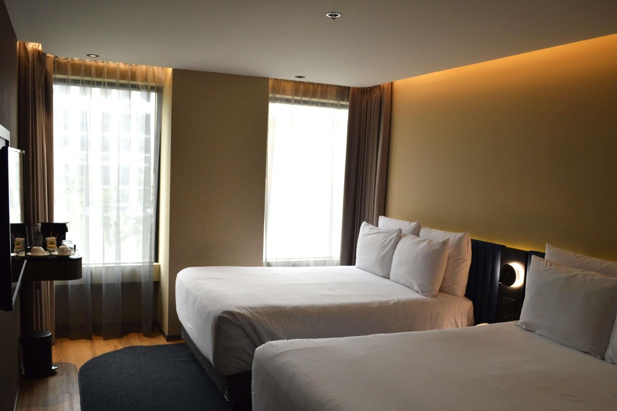 A hotel room with two beds, a bedside table, a lamp, and a window with curtains.