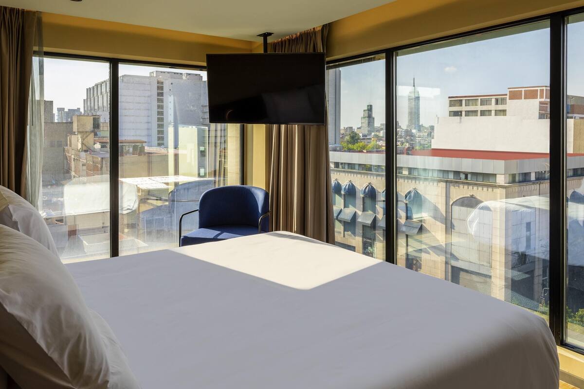 Una habitación de hotel con una cama grande, un televisor montado en la pared, una silla y un balcón con vista a la ciudad.