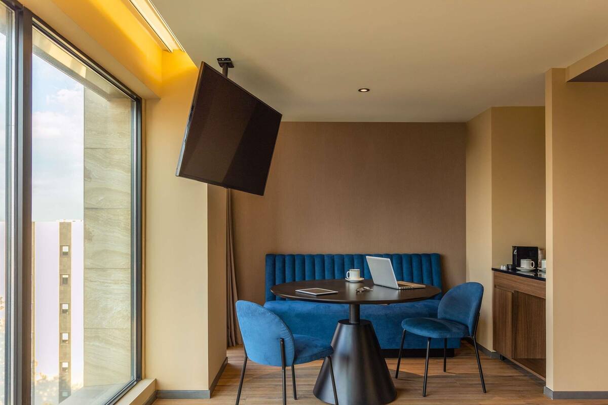 Una habitación moderna con una mesa redonda, sillas azules, un sofá, un televisor y una ventana con vistas a edificios.