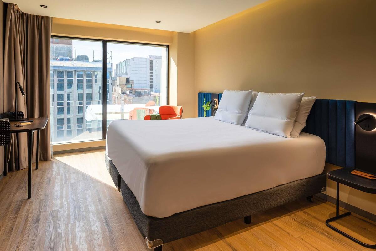 A hotel room with a large bed, a desk, a chair, and a view of the city through a large window.