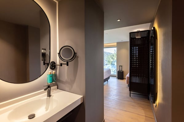 A modern bathroom with a large mirror, a round mirror, and a white sink. There is a bed and a suitcase in the adjacent room.