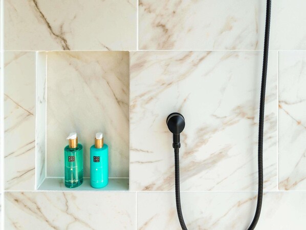 Un baño con pared de mármol, cabezal de ducha y dos botellas de jabón líquido.