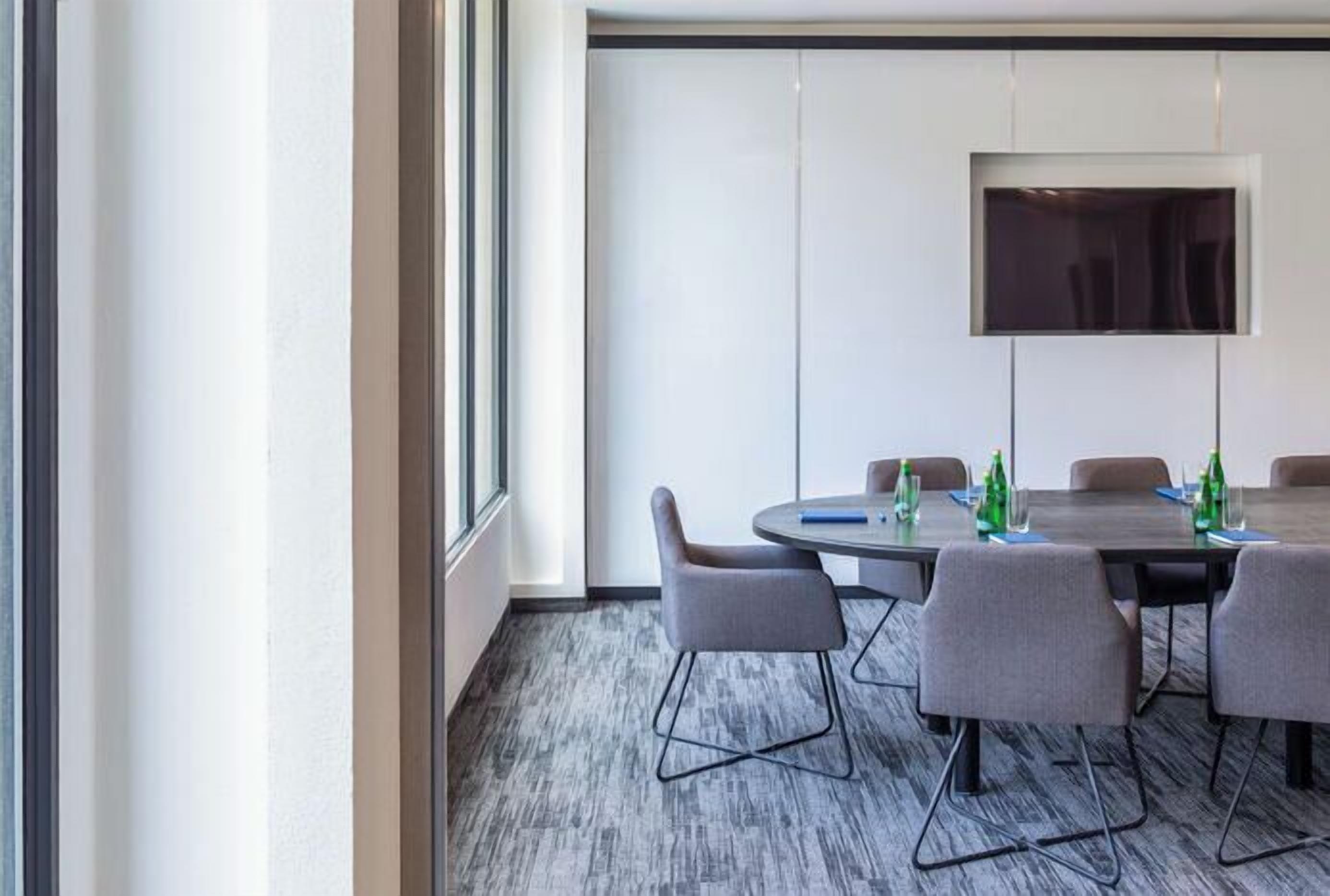 Ein Konferenzraum mit einem großen Tisch, grauen Stühlen, einem wandmontierten Flachbildfernseher und einem Fenster, das natürliches Licht hereinlässt.