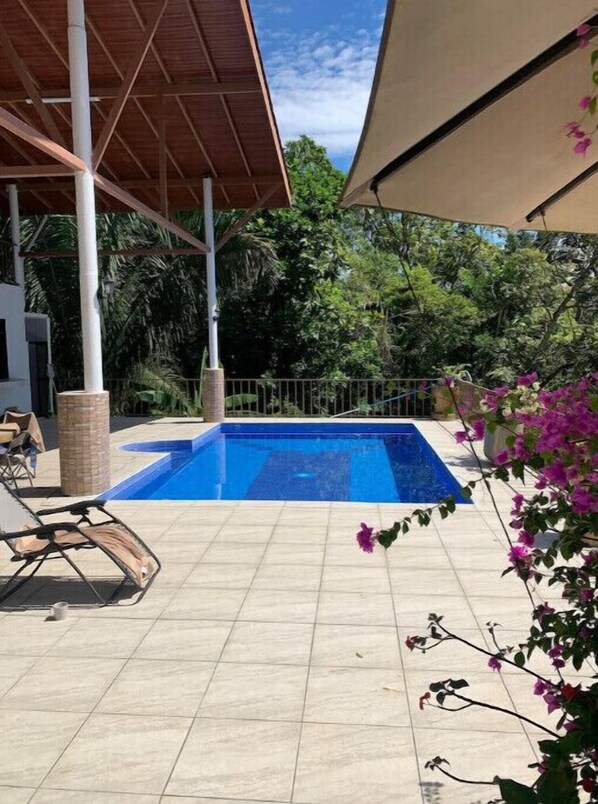 A swimming pool with a tiled deck, surrounded by greenery and a covered seating area.
