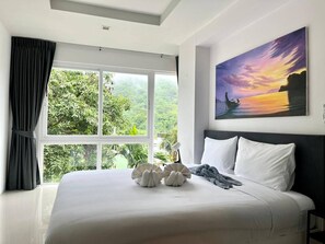 Une chambre avec un grand lit, un tableau représentant un coucher de soleil et une vue sur la verdure à l’extérieur.