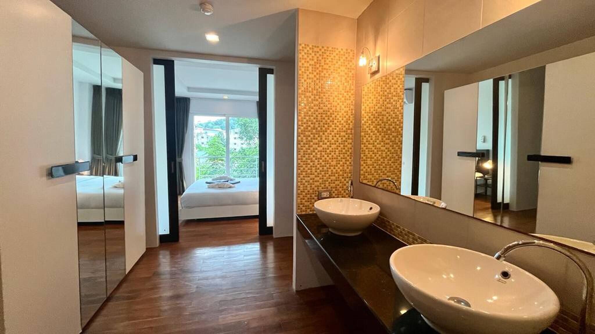 Ein modernes Badezimmer mit zwei Waschbecken, einem großen Spiegel und Blick auf ein Schlafzimmer mit Bett und Vorhängen.