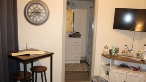 Una habitación con un escritorio de madera, dos taburetes, un reloj, un televisor, un mueble pequeño con diversos objetos y un cesto de basura.