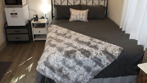 Een net opgemaakt bed met een grijze deken, een kussen met de tekst 'HAPPY PLACE', een nachtkastje met een lamp en een magnetron op een klein kastje.