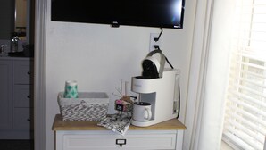 Een kleine, netjes ingerichte kamer met een wit bureau, een wandgemonteerde flatscreen-tv, een koffieapparaat en een raam met zonwering.