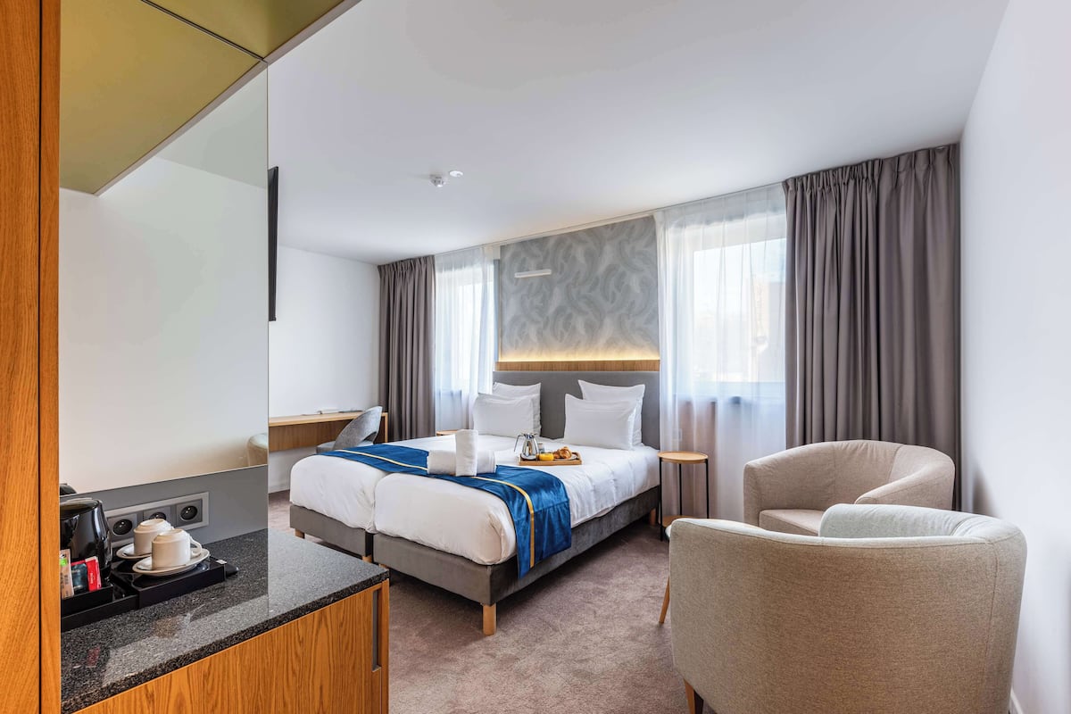 A hotel room with a bed, a chair, a desk with a coffee maker, and a window with curtains.