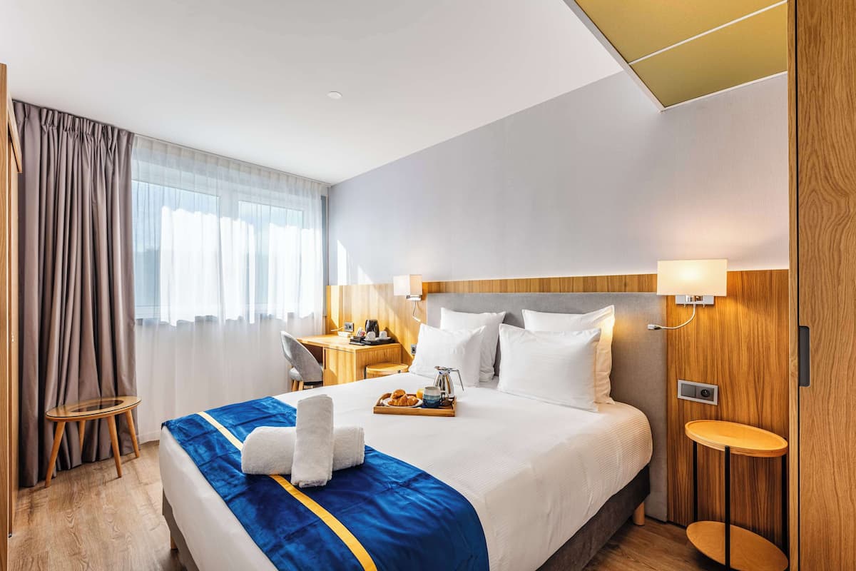 A hotel room with a large bed, a desk, a chair, a small table, and a window with curtains.