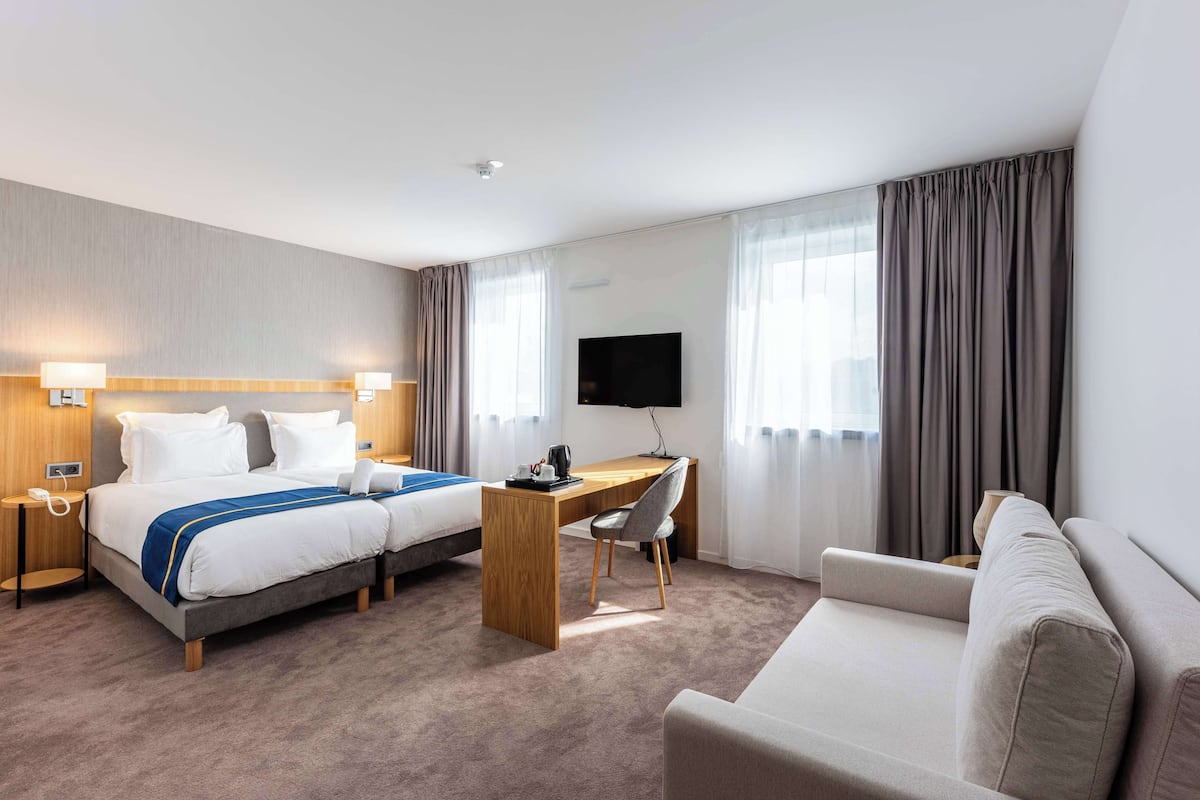 A hotel room with a bed, a desk, a chair, a sofa, a TV, and curtains.
