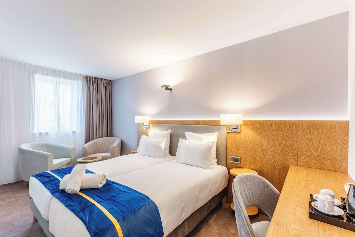 A hotel room with a large bed, two bedside tables, a desk with a chair, and a window with curtains.