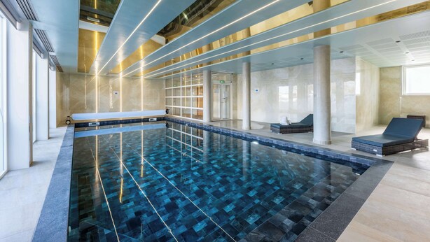 A modern indoor swimming pool with a clear blue water design, surrounded by lounge areas and a glass ceiling. - Brit Hotel Privilège Paris - Rosny (Rosny-sous-Bois)