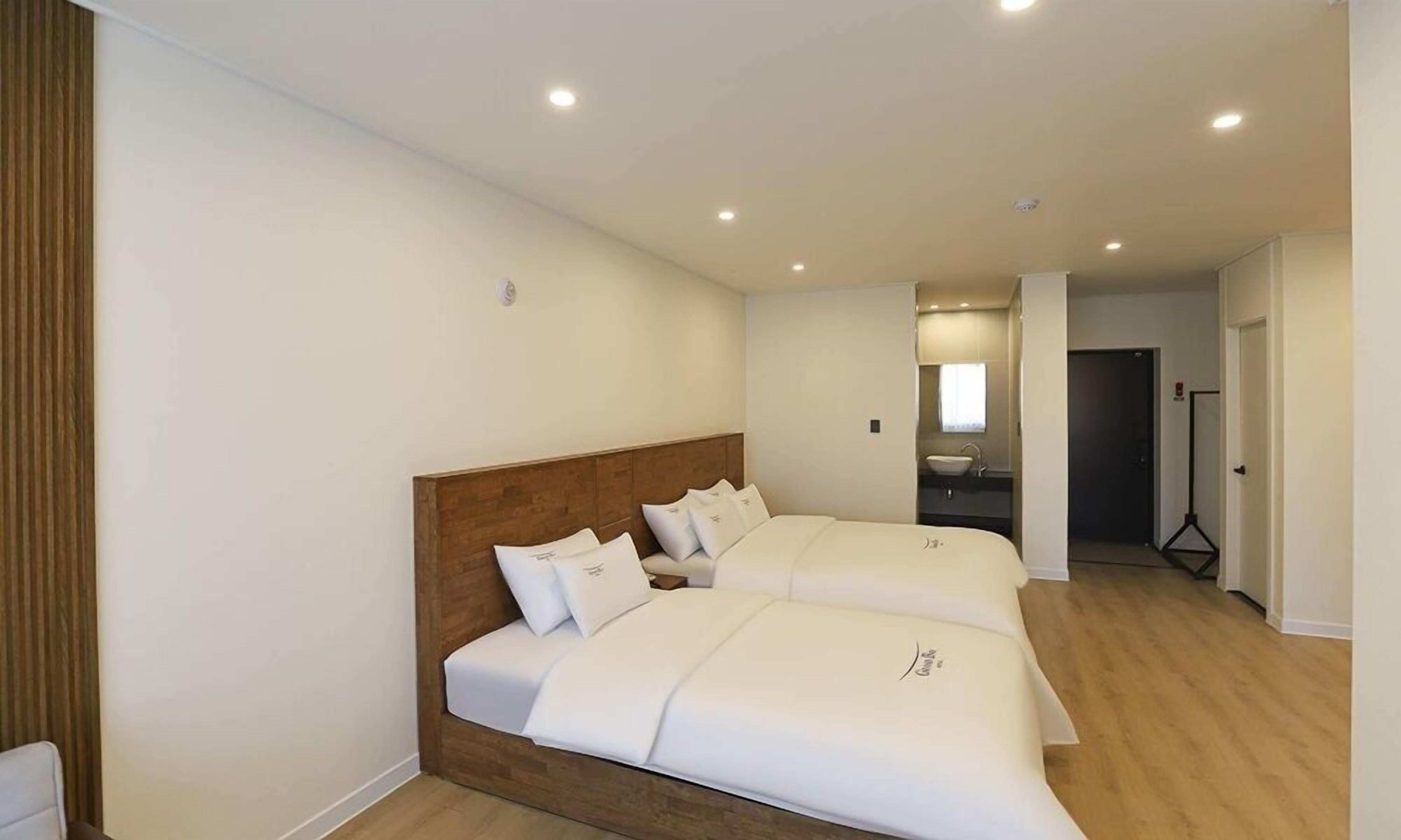 A hotel room with two beds, a wooden headboard, a bathroom with a sink and mirror, and a hallway leading to another room.