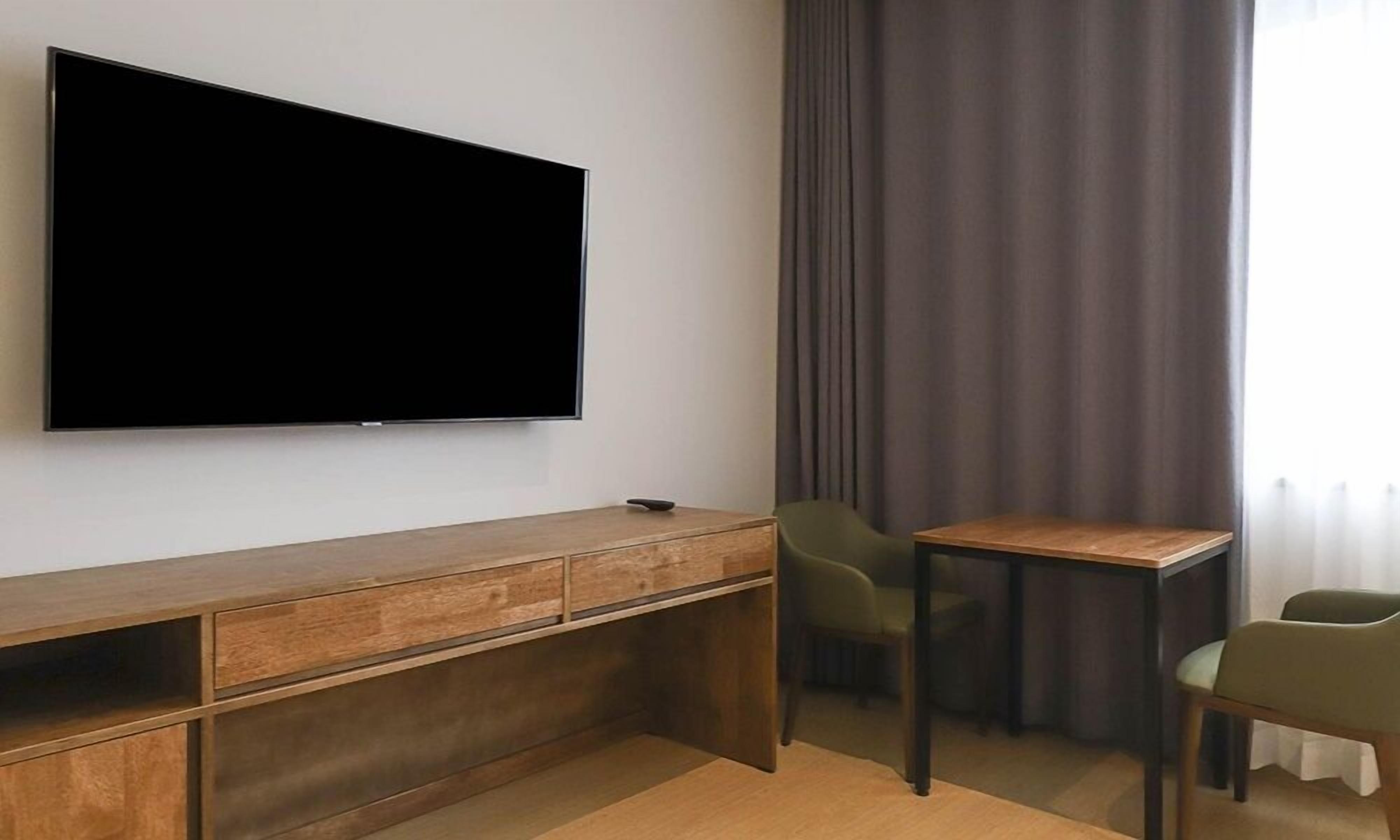 Quarto moderno com uma grande TV de tela plana na parede, uma escrivaninha de madeira com cadeira e uma janela com cortinas.