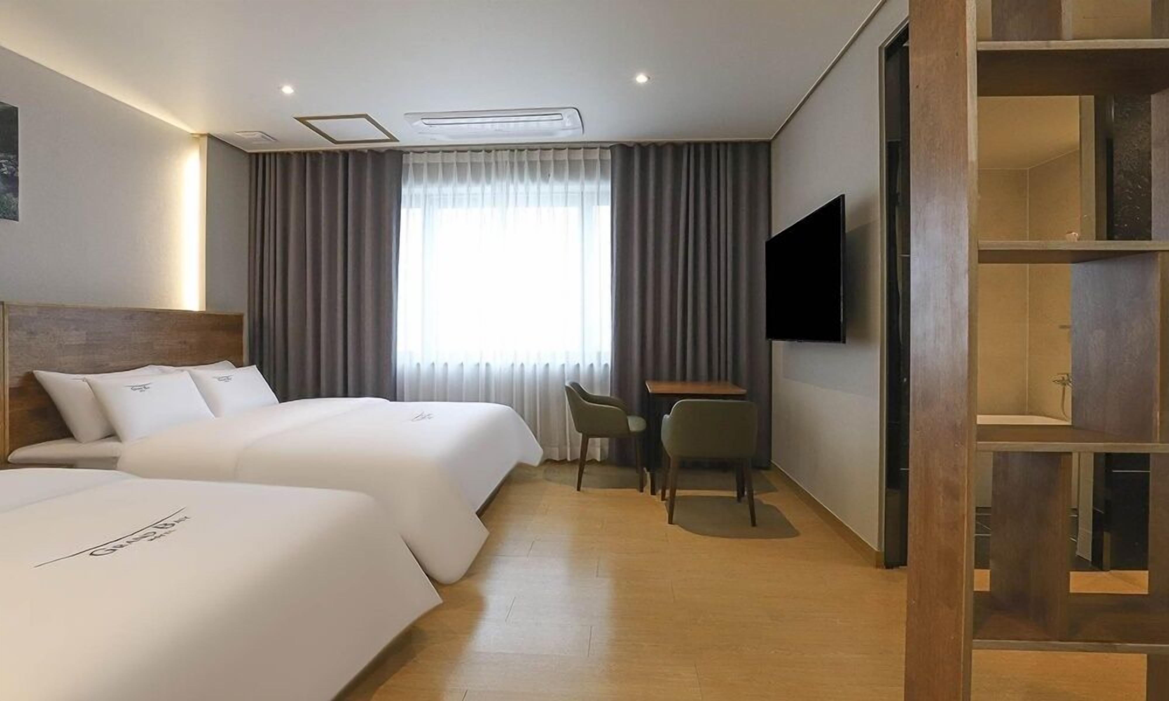 Habitación de hotel con dos camas, un escritorio, una silla, un televisor y un ventanal grande con cortinas.